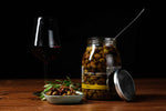 Jar of Taggiasca olives in extra virgin olive oil, small dish of olives, and glass of red wine on wooden table, gourmet Italian food from Nostrale Gourmet.